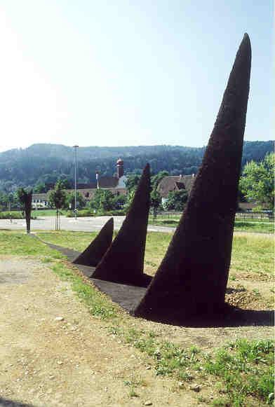 Monika Kaminska, Carouge GE
(Geschenk der Nordostschweizerischen Kraftwerke (NOK) AG an die Brown-Boveri&Co (BBC) zum 100-Jahr-Jubiläum)

Eine Form verschwindet im Boden. Die Dynamik der Bewegung hält an, setzt sich im Boden entlang einer imgaginären Linie fort und tritt wieder zutage in Form von drei Spitzen. Die formale Einfachtheit täuscht über die Komplexität des schöpferischen Prozesses hinweg, dessen Ergebnis diese Skulptur ist. Durch den Einbezug der geopraphischen  und symbolischen Gegenheiten des Standorts setzt sich das Werk in einem belebten Raum durch und schafft eine Verbindung zwischen alten und neuen Stadtquartieren. Ob dem Betrachter nun eher die Form oder die Aussage des Kunstwerkes wichtig ist – sein Blick verweilt, denn die Uebereinstimmung von Skulptur und Umgebung ist unmittelbar ersichtlich. 

Material: Ferrozement