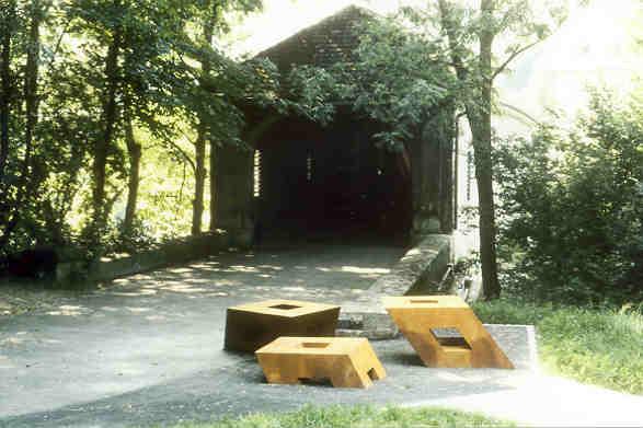 Peter Hächler, Lenzburg
(gestiftet durch die Gemeinde Neuenhof und Spenden Privater)

Es ist still geworden um die Holzbrücke, sie kommt sich verwaist und nutzlos vor, so auch die Mauern, die einst als Leitplanken den Verkehr ins enge Brückentor einfädelten, oder umgekehrt wieder der Landstrasse überliessen. Die drei gusseisernen Brocken, auch nutzlos, wie vom letzten schweren Fuhrwerk, das die Brücke passierte, gefallen und liegen gelassen, leisten der Brücke nun Gesellschaft, versuchen zu ihr zu gehören, mit ihr Ausdruck melancholischer Erinnerung an andere Zeiten.
Absicht ist, die stimmungsvolle räumliche Situation des Orts zu respektieren, diesen aber mit einem Akzent, mit einem unerwarteten „Ereignis“ in seiner Stimmung, seinem Ausdruck zu aktivieren..


Material: Eisenguss