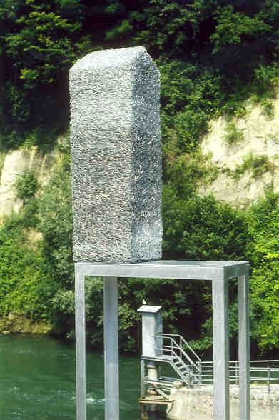 Lucie Schenker, St. Gallen
(gestiftet durch Coop und Ankauf Kulturwegstiftung)

Für ihr dreiteiliges Konzept hat Lucie Schenker die Holzbrücke, eine Mauer und das Pegelhäuschen als „Vorbilder“ ausgewählt. Auf einem Sockel wird dem Spaziergänger die kopierte Wirklichkeit in Modellform präsentiert. Im angepassten Massstab fordert die Künstlerin den Vorbeigehenden auf, seine eigens geschaffene Umwelt visuell wahrzunehmen. Das Modell hat anstelle einer zweckerfüllenden seine semiotische Funktion. 
Lucie Schenker verschlauft verzinkten Eisendraht so dicht ineinander bis aus dem filigranen Metall die kubische For des Vorbildes entsteht. Trotz der formalen Aehnlichkeit mit jener konstruierten Architektur ist der eindrückliche Entsehtungsprozess der Skulpturen mit organischem Wachstum zu vergleichen.

Material: Eisendraht verzinkt