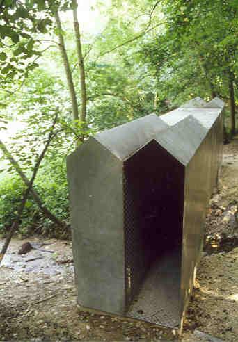 Kurt Sigrist, Sarnen
(gestiftet durch die Stadt Baden)

Zwei Einsiedeleien nennt Kurt Sigrist seine doppelte Metallbehausung. Eine Behausung, langgestreckt, korridorartig, nur einseitig geöffnet, nicht besonders einladend, aber doch Neugier erweckend, so empfängt das Metallgehäuse die Betrachter. Das Werk existiert seit 1985, aber hier am Krummbach, dem Grenzbach zwischen Baden und Neuenhof, hat der Künstler endlich einen treffenden Standort gefunden. Es verbindet brückenartig die beiden Ufer, aber nur optisch, nicht real. Und wer weitergeht, stellt sich in der Grossüberbauung Webermühle ähnliche Situtationen vor: Wohnung an Wohnung, mit Geräuschen durch die Wände. Jede und jeder lebt für sich, aber dem andern ausgesetzt.

Material:  Anticorodal-Riffelblech