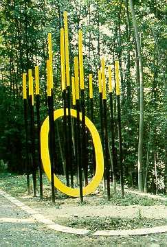 Gillian White, Leibstadt
(gestiftet durch Schweizerische Kreditanstalt Baden)

Gillian White lässt sich durch den Ort – eine Lichtung in einem jungen Wald inspirieren. Mit den schmalen Stangen nimmt sie das dunkle Stammgewirr auf und akzentuiert das Umgebungsbild. Das leuchtende Gelb der Stangenspitzen erinnert an  die Sonnenlicht aufblitzenden Laubpartien und Baumspitzen. Die gelbe Ellipse in der Werkmitte nimmt die Erscheinung der Lichtkreise auf, wie sie an strahlenden Sonnentagen um die Stämme entstehen.

Material: Stahl bemalt, Kalksteine