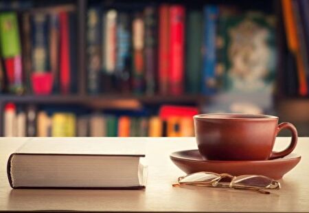 Buch, dampfender Kaffee und Lesebrille liegen auf einem Tisch vor einem Büchergestell