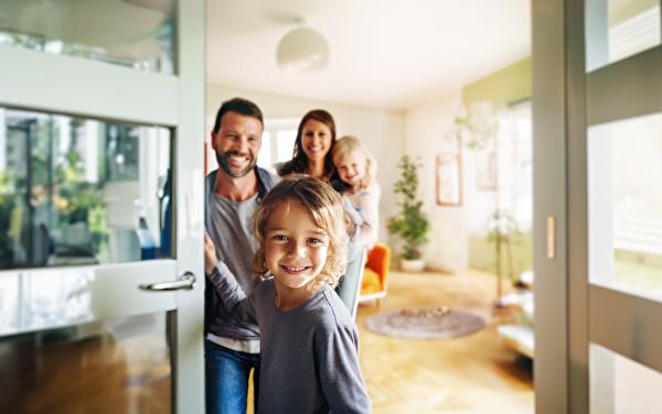 Symbolbild einer Familie, die jemanden an der Tür empfängt. Das kleine Mädchen er Familie öffnet die Türe, die anderen (Elstern und kleinere Schwester) stehen lachend im Hintergrund