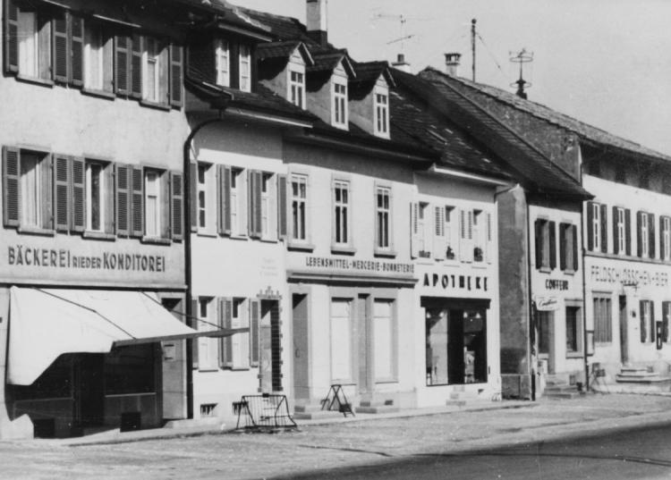 V.l.n.r.: Bäckerei und Konditorei Rieder, Lebensmittelgeschäft, Apotheke Leupin, Coiffeur, (Warteck) Feldschlösschen Bier

Datierung: keine Angaben