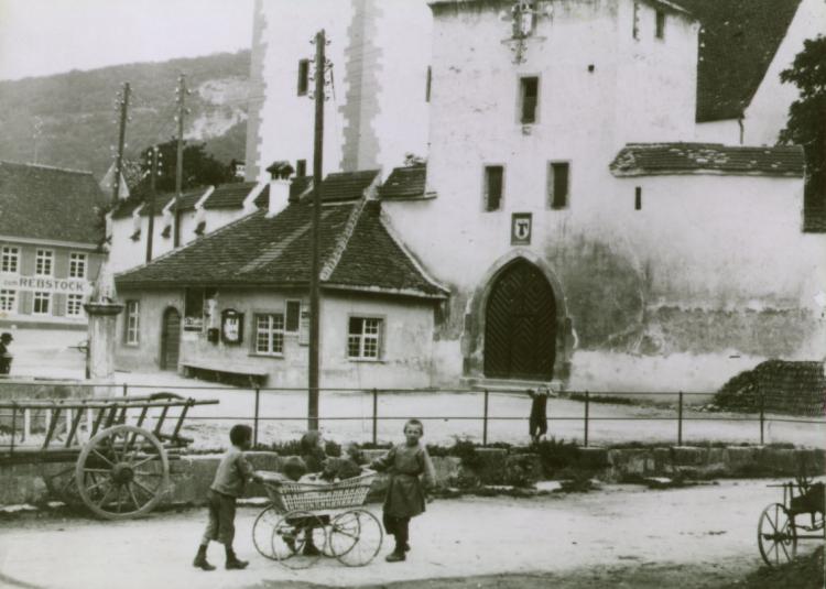 Spielende Kinder vor der Kirche St. Arbogast.
An der Kirchmauer befindet sich das Wachthaus.

Der Dorfbach ist hier noch offen und durch ein Geländer gesichert.


