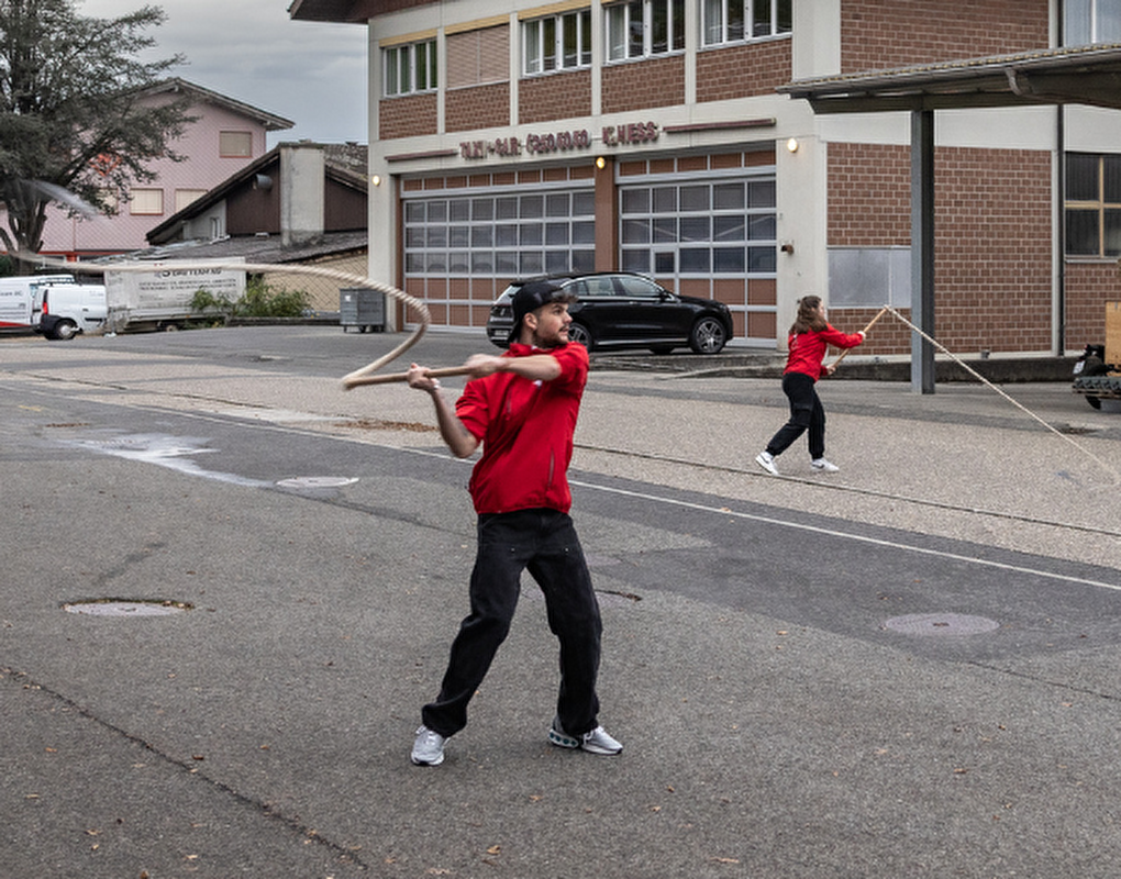 Vier Personen, alle in roten Jacken, stehen auf einer Strasse und praktizieren den traditionellen Brauch des Geisslechlöpfe. Sie schwingen lange, flexible Ruten und führen eine koordinierte Bewegung aus. Die Szene spielt sich an einem bewölkten Tag ab, im Hintergrund sind geparkte Autos und Gebäude zu sehen, darunter ein grosses Gebäude mit einer Garage.