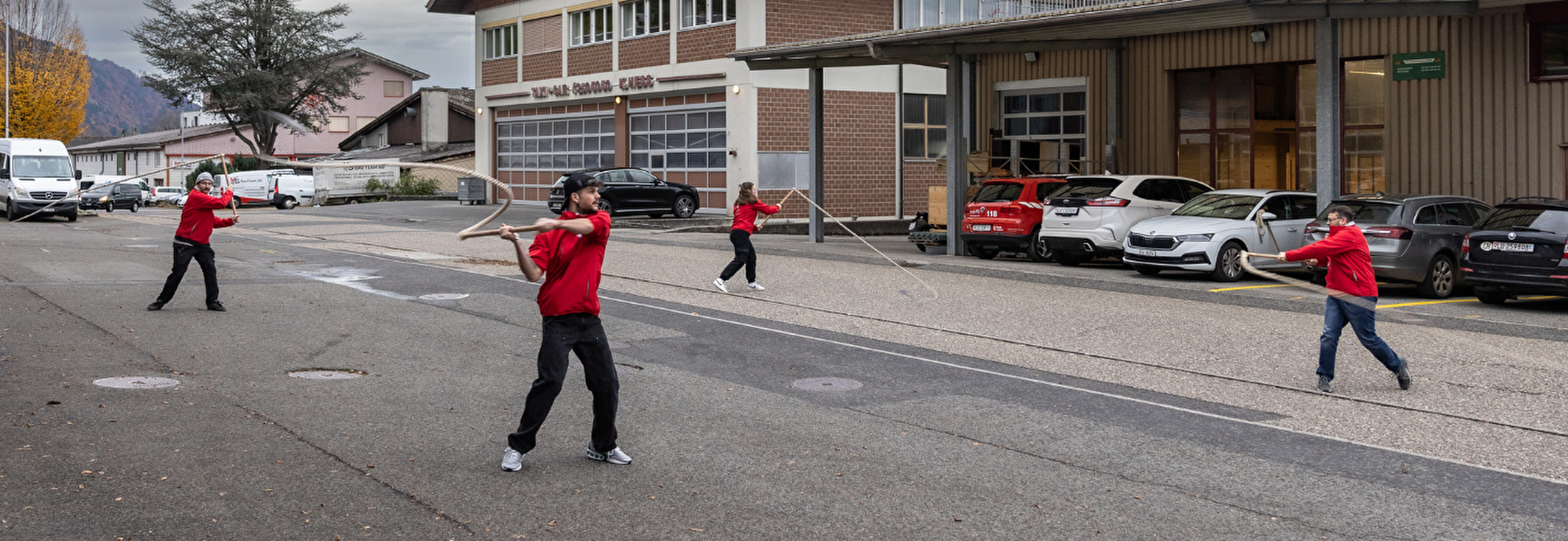 Vier Personen, alle in roten Jacken, stehen auf einer Strasse und praktizieren den traditionellen Brauch des Geisslechlöpfe. Sie schwingen lange, flexible Ruten und führen eine koordinierte Bewegung aus. Die Szene spielt sich an einem bewölkten Tag ab, im Hintergrund sind geparkte Autos und Gebäude zu sehen, darunter ein grosses Gebäude mit einer Garage.