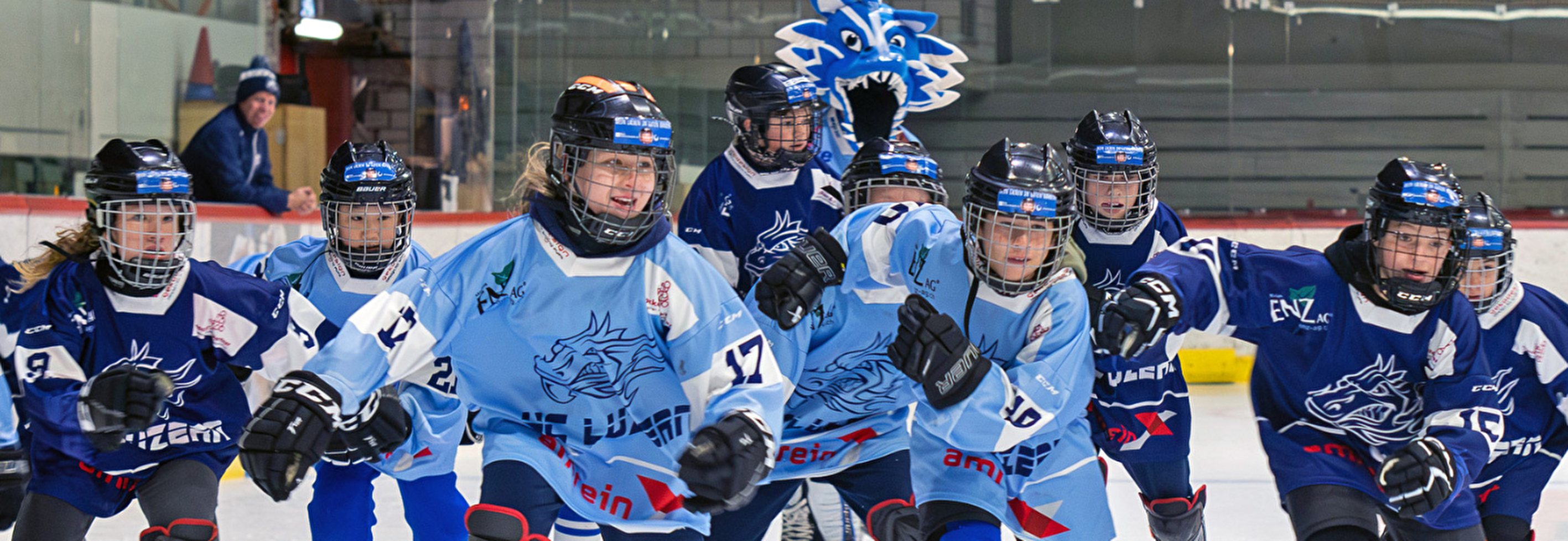 Gruppenbild von jungen Eishockeyspielerinnen des Hockey Club Luzern in Aktion auf der Eisfläche. Die Spielerinnen tragen blaue Trikots mit dem Logo des Clubs und Schutzkleidung. Einige tragen hellblaue Trikots, während andere dunkelblaue Trikots tragen. Im Hintergrund ist eine Person in Zivilkleidung zu sehen, die hinter der Bande steht. Das Bild zeigt die Dynamik und den Teamgeist des Clubs, der kürzlich den Sportpreis 2025 gewonnen hat. Quelle: Hockey Club Luzern