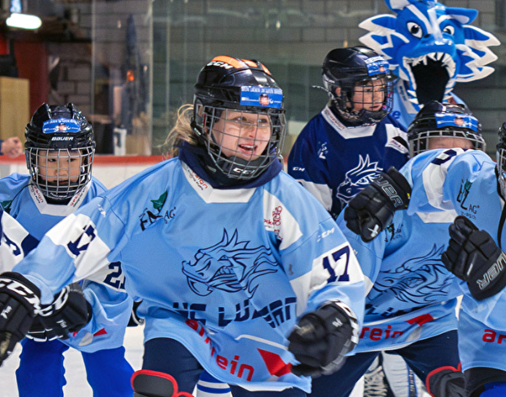Gruppenbild von jungen Eishockeyspielerinnen des Hockey Club Luzern in Aktion auf der Eisfläche. Die Spielerinnen tragen blaue Trikots mit dem Logo des Clubs und Schutzkleidung. Einige tragen hellblaue Trikots, während andere dunkelblaue Trikots tragen. Im Hintergrund ist eine Person in Zivilkleidung zu sehen, die hinter der Bande steht. Das Bild zeigt die Dynamik und den Teamgeist des Clubs, der kürzlich den Sportpreis 2025 gewonnen hat. Quelle: Hockey Club Luzern