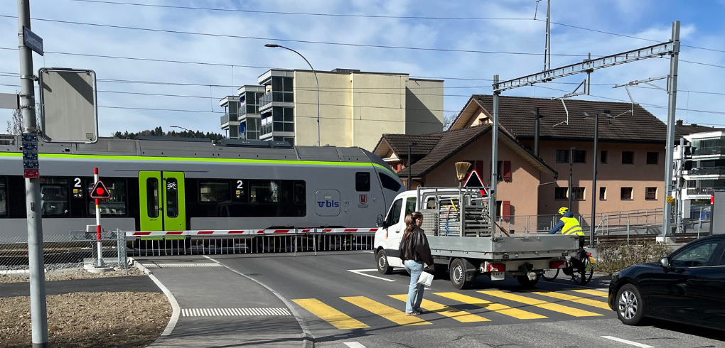 Bahnübergang in Littau mit gesenkter Schranke und durchfahrendem Zug, welcher Grau und Grün lackiert ist. Vor der Bahnschranke steht ein weisser Pritschenwagen, eine junge Frau überquert einen Fussgängerstreifen und ein Velofahrer mit einer Leuchtweste wartet darauf, den Bahnübergang überqueren zu dürfen.