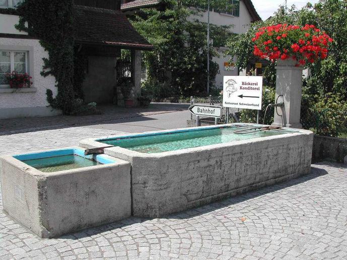 Hier trennen sich Bahnhof- und Dorfstrasse und bilden einen spitzen Winkel. Daher wohl der Name Spitz-Brunnen. Er trägt das Jahr 1870 und ist mit 3 Liter/Minute dem Leitungsnetz angeschlossen.