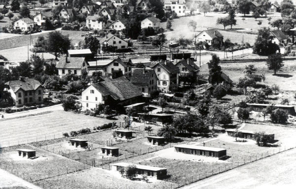 Foto um 1950
Die Häuser und die Hühnerfarm Baumann stehen unmittelbar oberhalb des mit Barrieren versehenen Bahnübergangs.