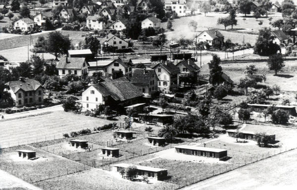 Foto um 1950
Die Häuser und die Hühnerfarm Baumann stehen unmittelbar oberhalb des mit Barrieren versehenen Bahnübergangs.