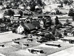 Foto um 1950
Die Häuser und die Hühnerfarm Baumann stehen unmittelbar oberhalb des mit Barrieren versehenen Bahnübergangs.