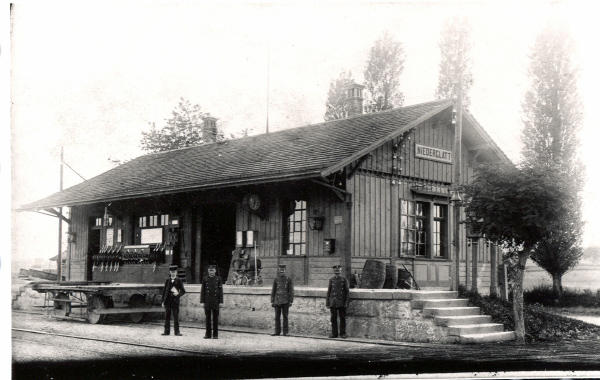 Aufnahme ca. 1905
<b>Das erste Stationsgebäude in Niederglatt</b> und die Bahnlinie Oerlikon - Niederglatt - Bülach wurden <b>im Jahre 1865 in Betrieb</b> genommen. Dieses Photo dürfte jedoch erst nach der Jahrhundertwende um 1900 entstanden sein. Das Personal bestand aus einem Vorstand, einem "Gehülfen" und zwei Stationswärtern . Es ist bemerkenswert, dass bereits ein Stellwerk vorhanden war.
Für den Bau des neuen Stellwerks im Jahre 1909 wurden viele Teile wiederverwendet. Aus dem Stationsgebäude entstand später der Schuppen.
An der Stirnfront, zwischen dem Fenster und der Stationsschrift hänqt die Glocke, womit die Passagiere jeweils zum Einsteigen aufgefordert wurden.
Mit dem Leiterwagen wurden die Güter zur Bahn gebracht oder dort abgeholt.