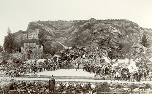 Schwarz-weiss-Fotografie mit einer grossen Bühne. Das Bühnenbild zeigt ein Bergpanorama und Burgen, auf der Bühne die aufgestellten Krieger in historischem Gewand, mit Spiessen bewaffnet. Im Vordergrund Zuschauer und das Orchster.
