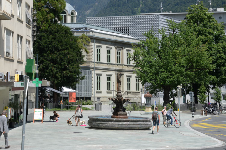 Der Postplatz auf der Seite Bahnhofstrasse mit dem seit 1880 sprudelnden Brunnen, dem Bündner Kunstmuseum mit den beiden Gebäuden Villa Planta und dem halbverdeckten, im Juni 2016 eröffneten, Erweiterungsbau dahinter.