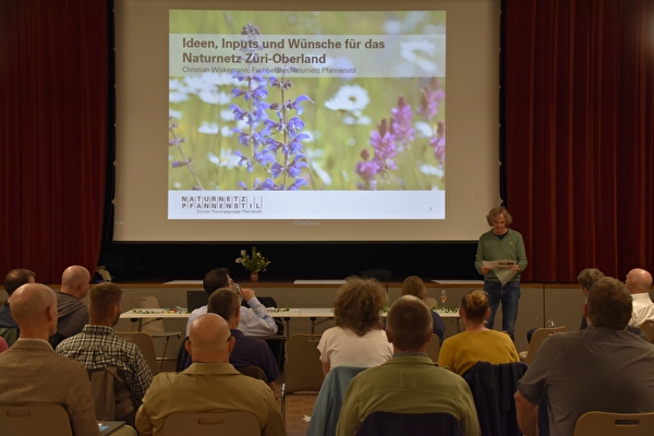 Foto von Personen in einem Saal mit Bühne und Projektion an der Leinwand