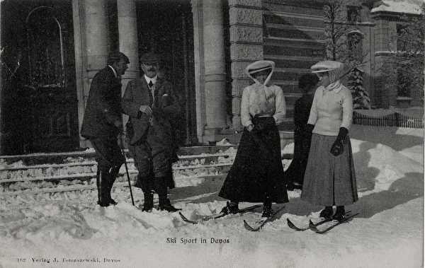 Zwei Damen mit mit Hut, Rock und Skiern an den Füssen sowie zwei Herren vor dem verschneiten Eingang des Postgebäudes ca. 1906