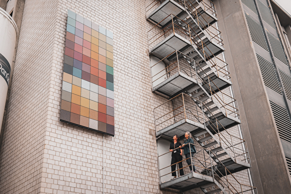 Zwei Frauen schauen von einer Treppe aus auf farbige Solarpanels an der Wand.