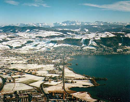 Pfeilgerade führt die Bahnlinie ins Zentrum von Zug. Im Vordergund das Quartier Riedmatt. Hinter dem Zugerberg das Panorama der Glarner Alpen.