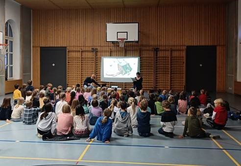 Kinder sitzen in der Turnhalle am Boden und schauen den Darbietungen zu