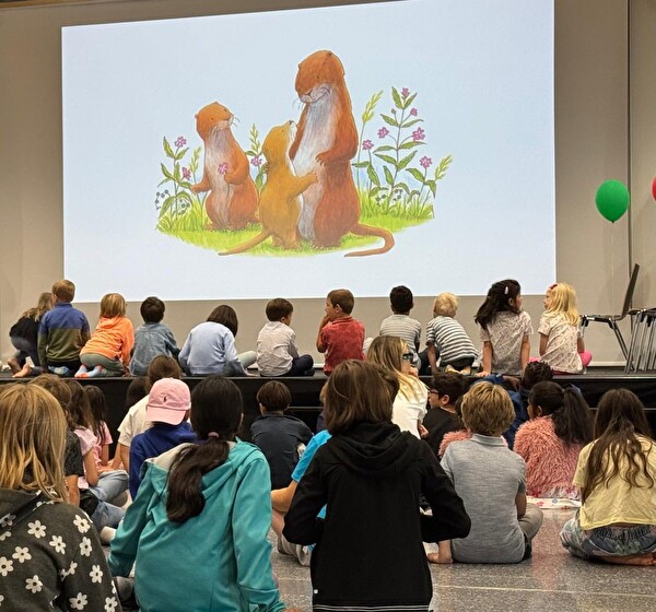 Kinder sitzen vor der Bühne, schauen auf die Leinwand, sehen eine Hamster Familie mit einem Kind