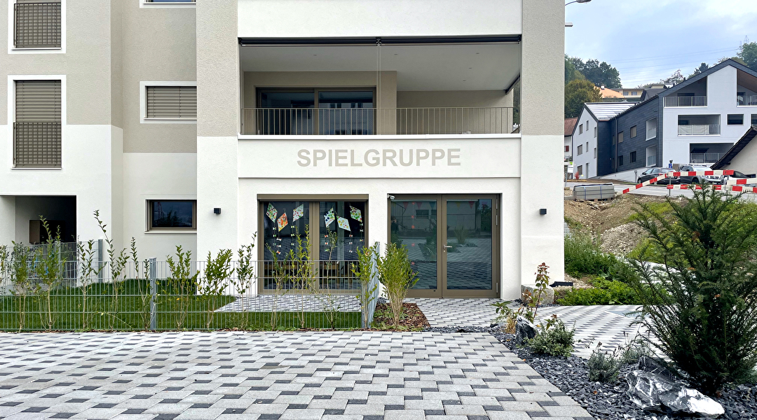 Front von modernem Mehrfamilienhaus. Oberhalb des Einganges steht Spielgruppe