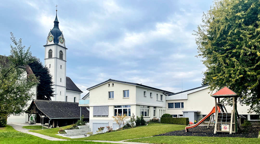 Kindergarten Pfarreiheim mit Spielplatz im Vordergrund und Kirche im Hintergrund