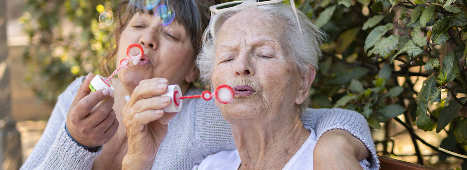 Zwei Seniorinnen sitzen auf einer Parkbank und machen Seifenblasen.
