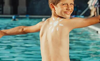 Ein Kind am Schwimmbecken Rand zwinkert uns zu und hat eine Zeichnung einer Sonne, mit Sonnencreme gemacht, auf dem Rücken.