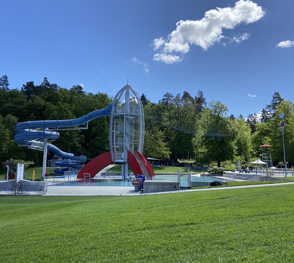 Freibad Kloten: Von der grünen Wiese aus mit Sicht auf die Rakete mit den Wasserrutschen und den Hardwald im Hintergrund.